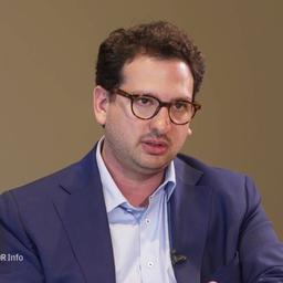 Ein Mann mit braunen Haaren, Hornbrille und blauen Anzug sitzt an einem Tisch.