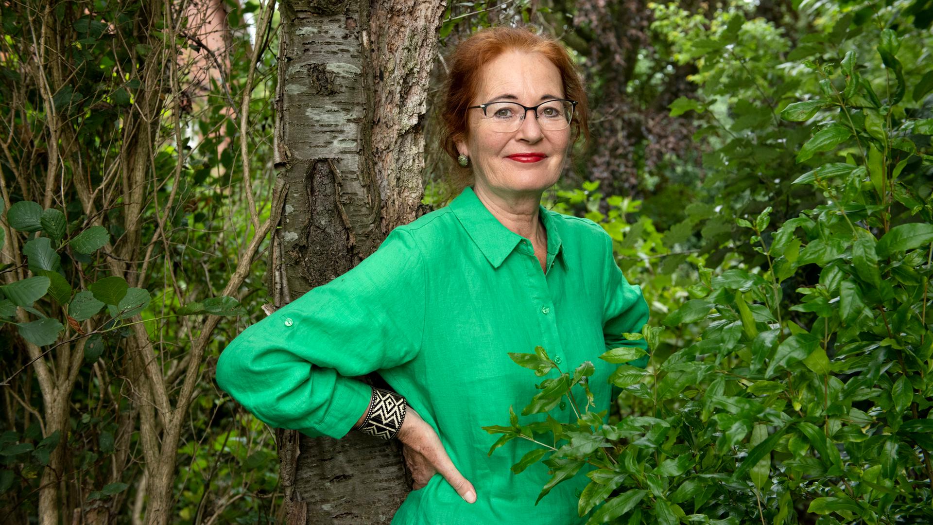 Eine Frau mit Brille und grüner Bluse lehnt an einem Baumstamm, umgeben von Grün | Eva Häberle