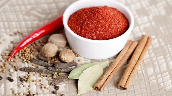 Eine Schale mit einer Gewürzmischung sowie Zimtstangen, Muskatnüsse, Langer Pfeffer, Lorbeerblätter und eine Chillischote.