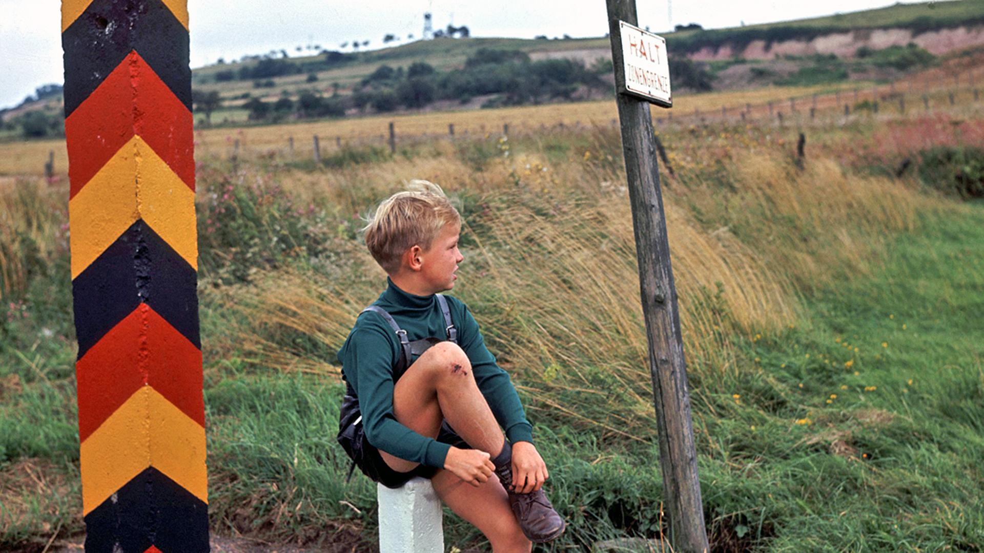 Ein Junge sitzt auf einem weißen Pfosten zwischen einem Grenzpfahl mit dem Wappen der DDR und einem Schild mit der Aufschrift 