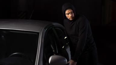 Ein schwarz gekleideter Mann bricht in ein Auto ein.