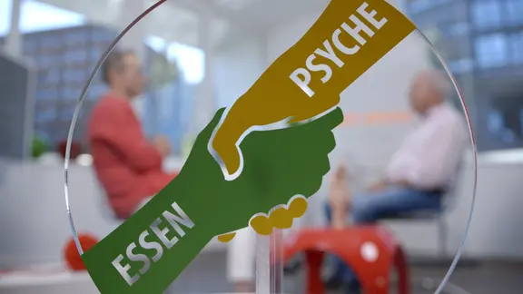 Eine halbtransparente Tafel zeigt den Händedruck von "Essen" und "Psyche", dahinter sitzen Doc Riedl und sein Patient. | NDR, Moritz Schwarz/Oliver Zydek Eine halbtransparente Tafel zeigt den Händedruck von "Essen" und "Psyche", dahinter sitzen Doc Riedl und sein Patient.