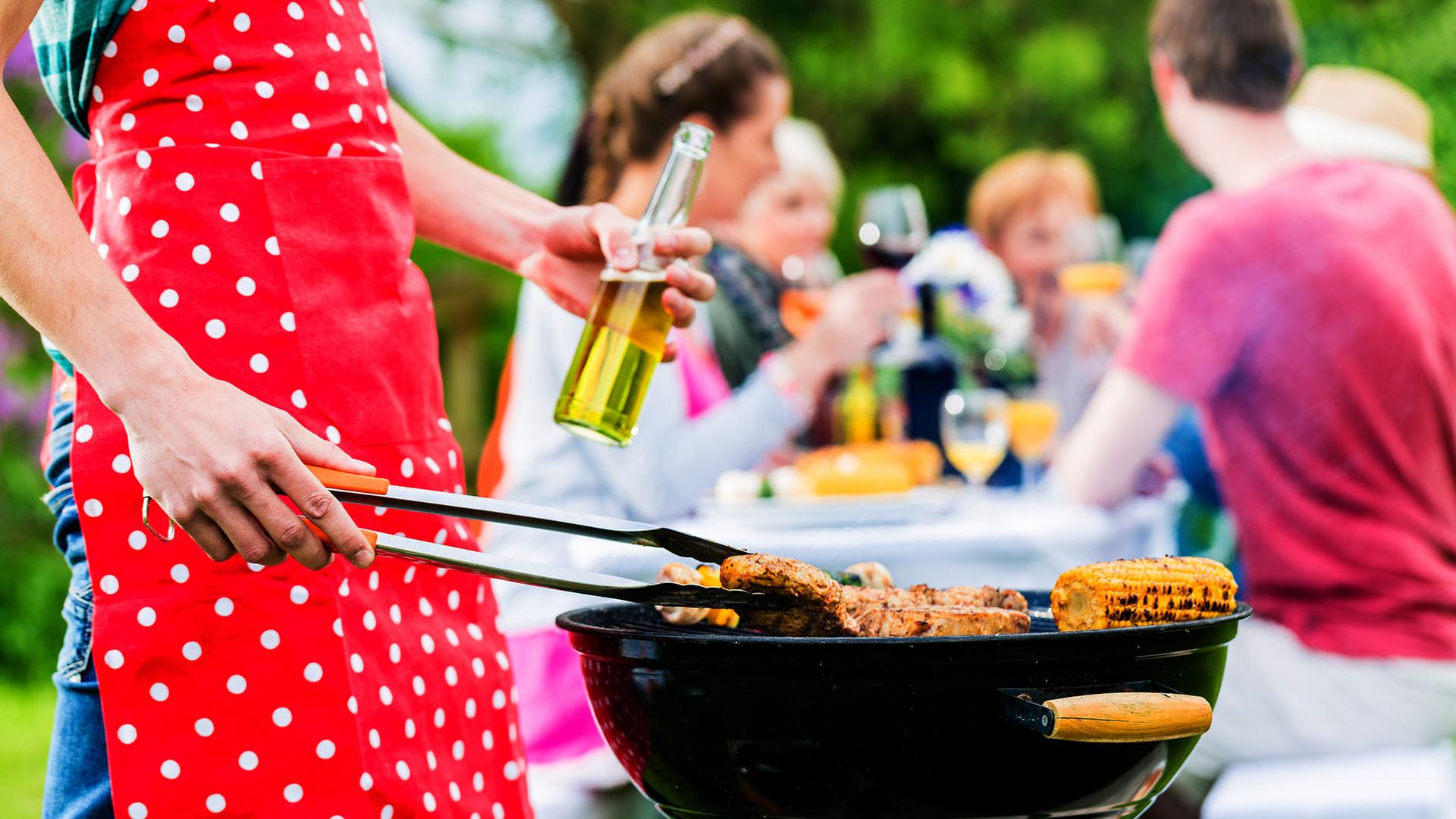 Person steht mit einer Schürze bekleidet am Grill, im Hintergund essen und trinken Leute. | Fotolia, Kzenon