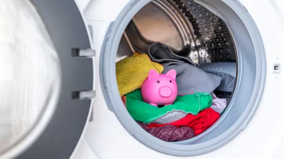 Blick in eine Waschmaschine, in der auf der Wäsche ein Sparschwein steht.