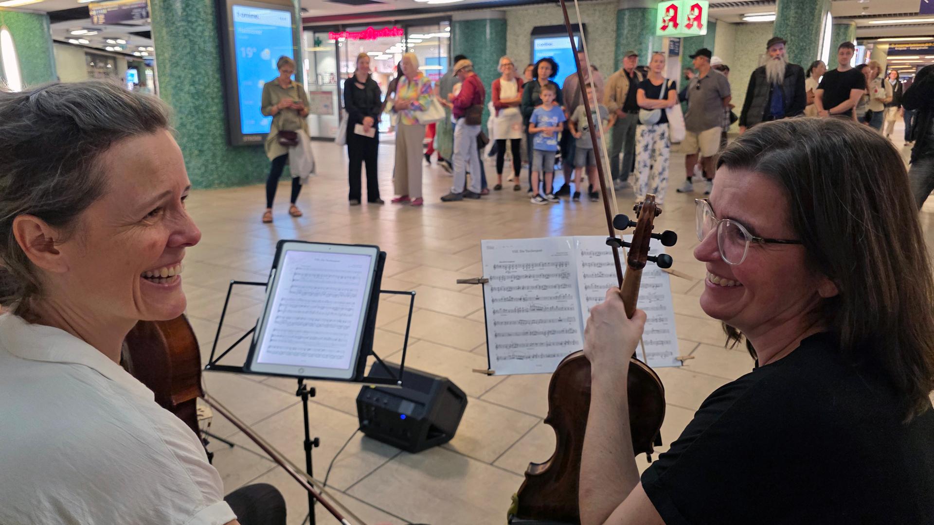 Zwei Musikerinnen sitzen mit ihren Instrumenten, einer Geige und einem Cello, vor Notenständern und lächeln sich gegenseitig an. Im Hintergrund stehen zahlreiche Menschen in einer Bahnhofshalle und beobachten das Duo.  | NDR, Foto: Agnes Bührig 