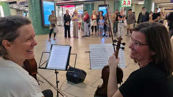 Zwei Musikerinnen sitzen mit ihren Instrumenten, einer Geige und einem Cello, vor Notenständern und lächeln sich gegenseitig an. Im Hintergrund stehen zahlreiche Menschen in einer Bahnhofshalle und beobachten das Duo. 