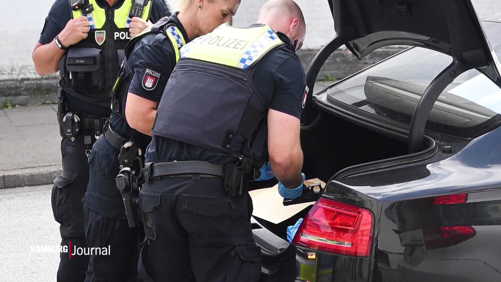 Mehrere Polizeibeamte durchsuchen einen Kofferraum eines schwarzen PKW; ein Polizist hält eine gefundene Pistole in der Hand. | Screenshot
