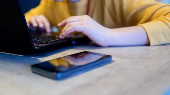 Ein Kind sitzt vor einem Laptop und einem Handy und schreibt auf der Tastatur.
