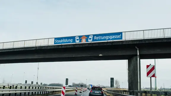 An dem Geländer einer Autobahnbrücke hängt ein Hinweisschild mit der Aufschrift Staubildung: Rettungsgasse.