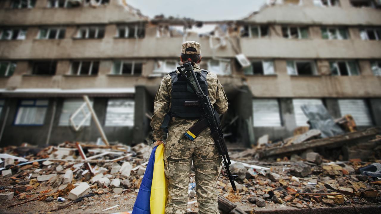 Ukrainische Soldatin mit ukrainischer Flagge in den Händen vor dem Hintergrund eines explodierten Hauses