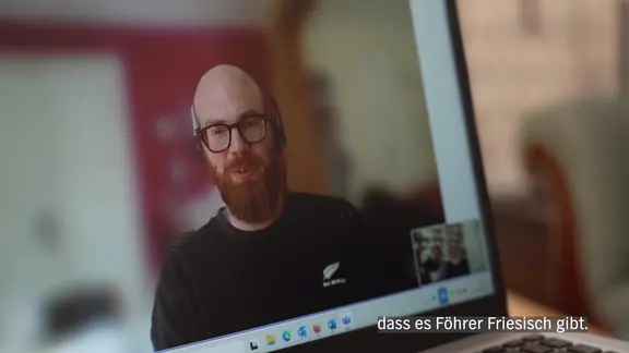 Ein Mann auf einem Computerbildschirm mit Glatze und Bart spricht.