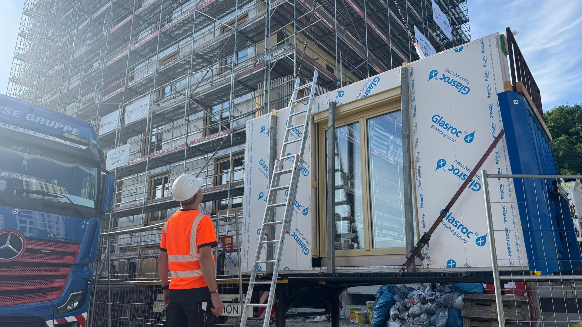 Ein Bauarbeiter steht vor einem fertigen Holz-Modul vor einem Hochhaus. | NDR, Sabine Hausherr