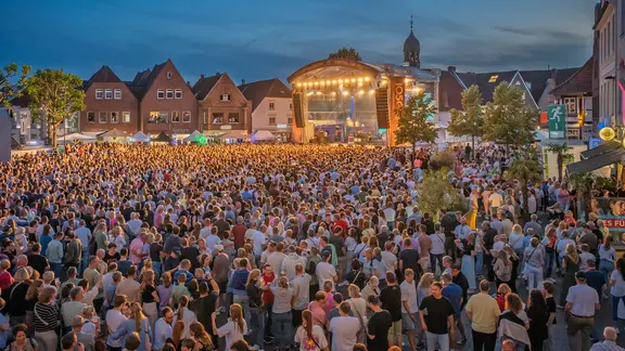 Eine Menschenmenge steht vor einer Bühne auf dem Marktplatz in Lingen. Es ist Abend.