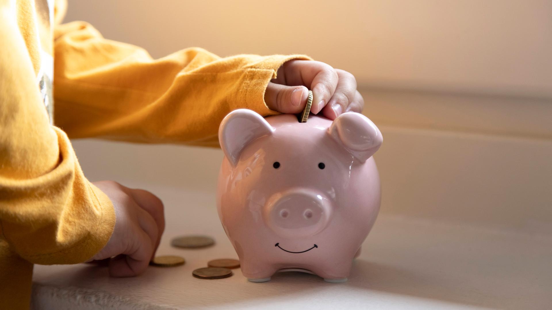 Ein Kind packt Kleingeld in ein Sparschwein. | IMAGO / Westend61